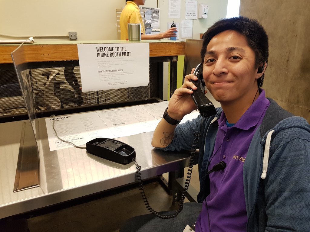A man sitting at table, using a landline phone and smiling at the camera. In front of the table there's a piece of paper that says "welcome to the phone booth pilot".