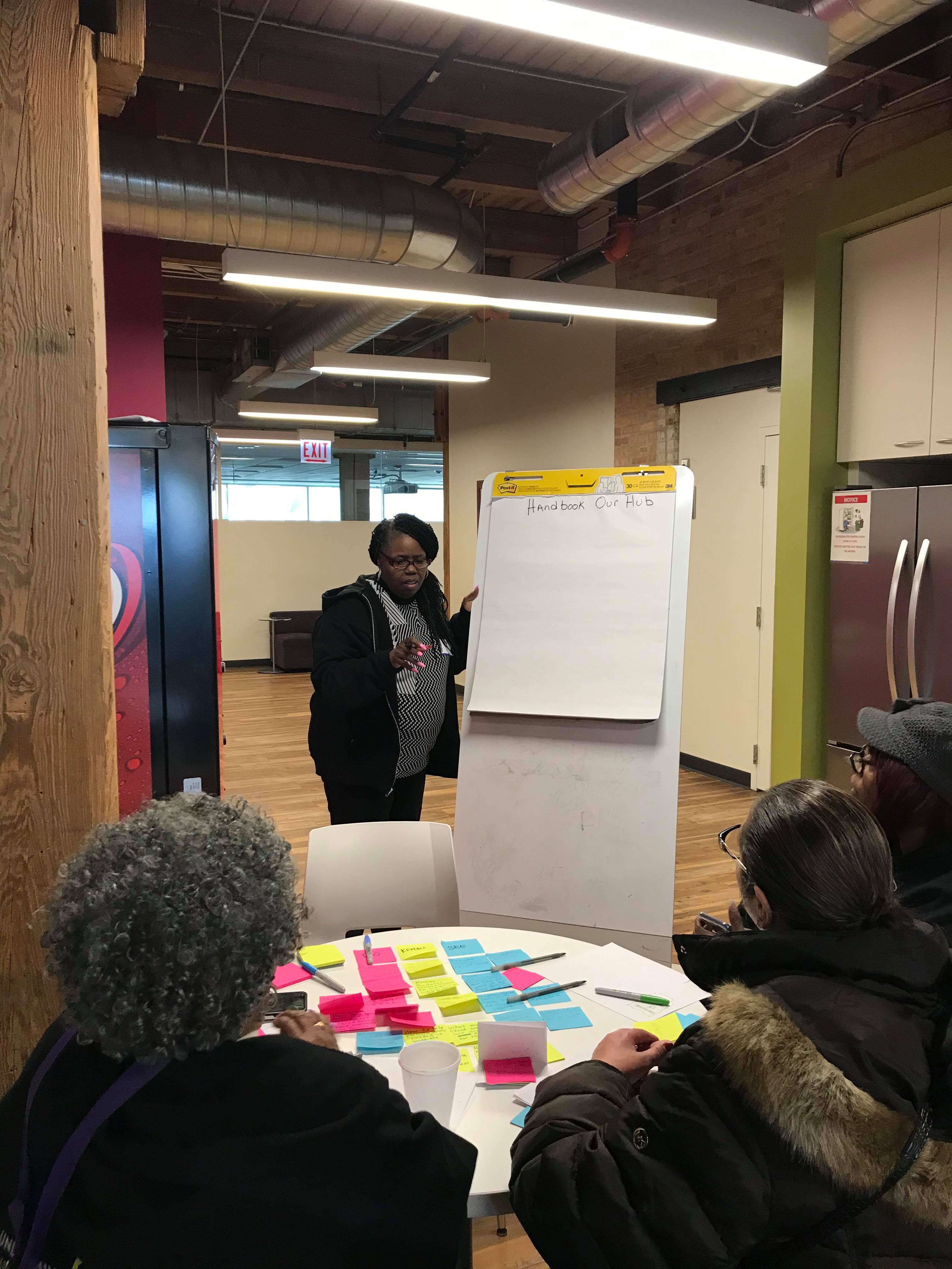 A woman stands in front of a two other women, with a large piece of paper on a board next to her and a pen in her hand. The piece of paper has been titled “Handbook, our hub”. Two women who are sitting at a small round table covered in post-it notes look at her.