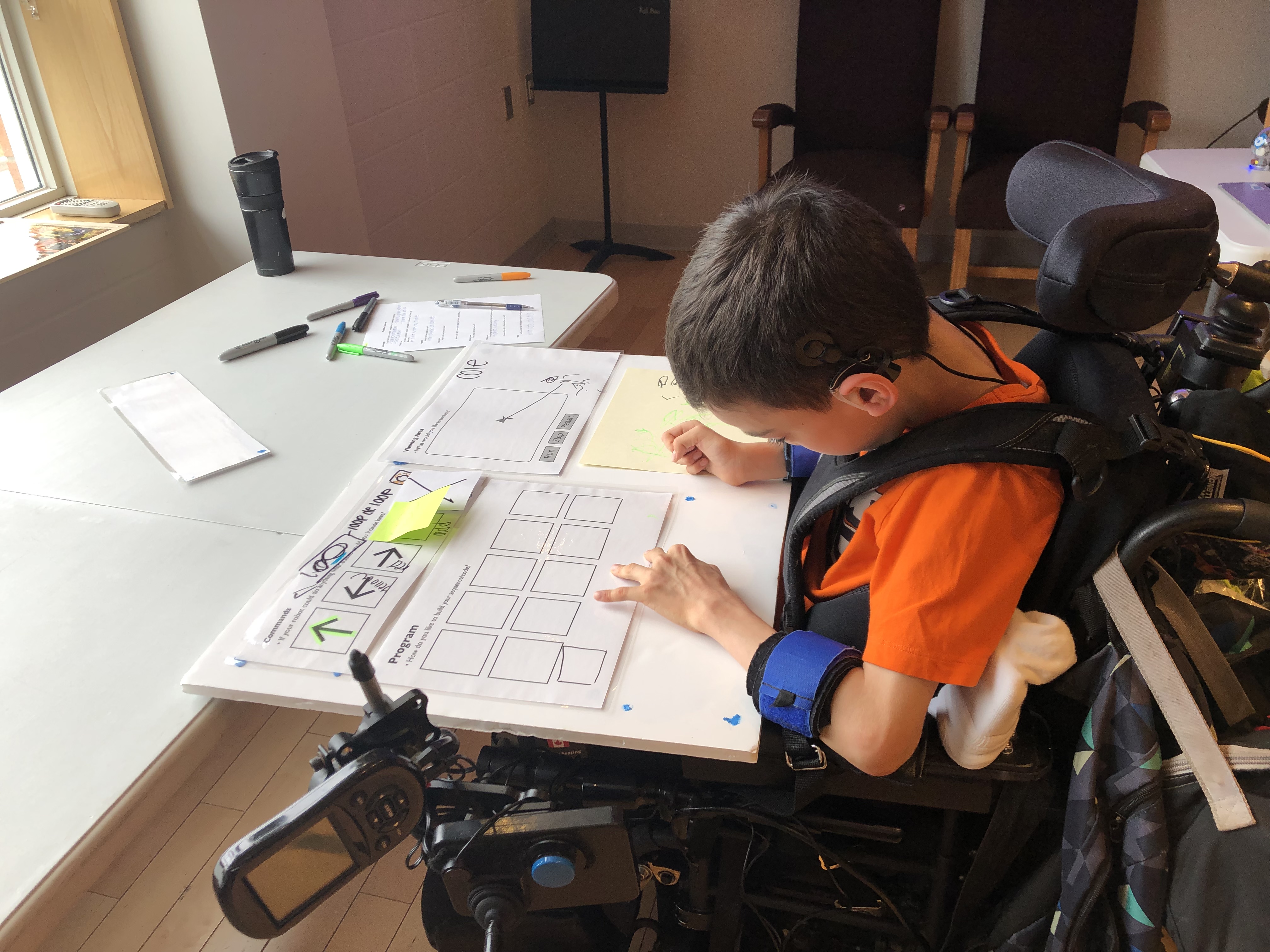 A student in a wheelchair is interacting with a large paper prototype of the C2LC coding environment.  