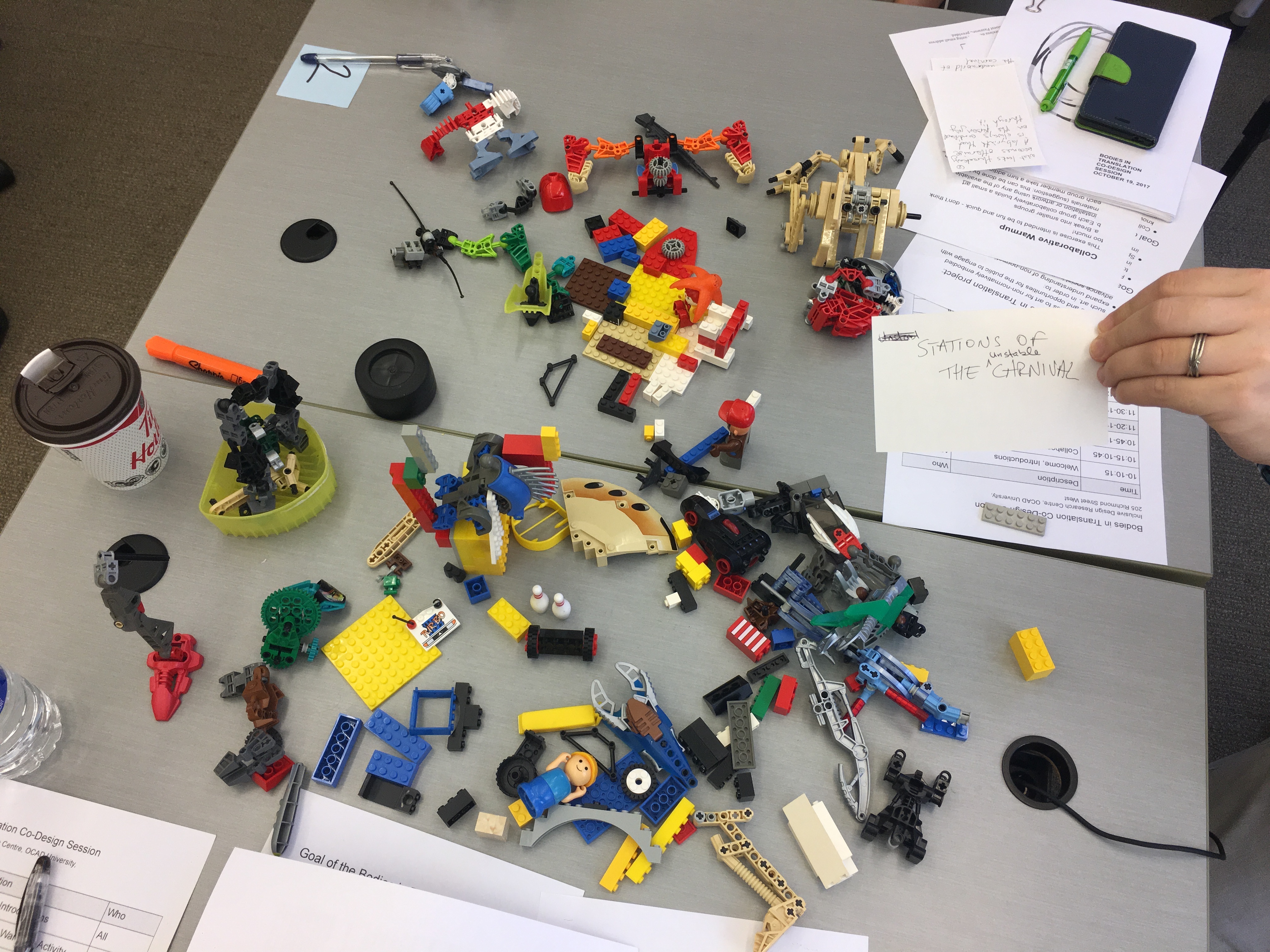 Photo of a desktop filled with various lego constructions. A hand can be seen on the right side holding up a note that says "Stations of the Unstable Carnival"
