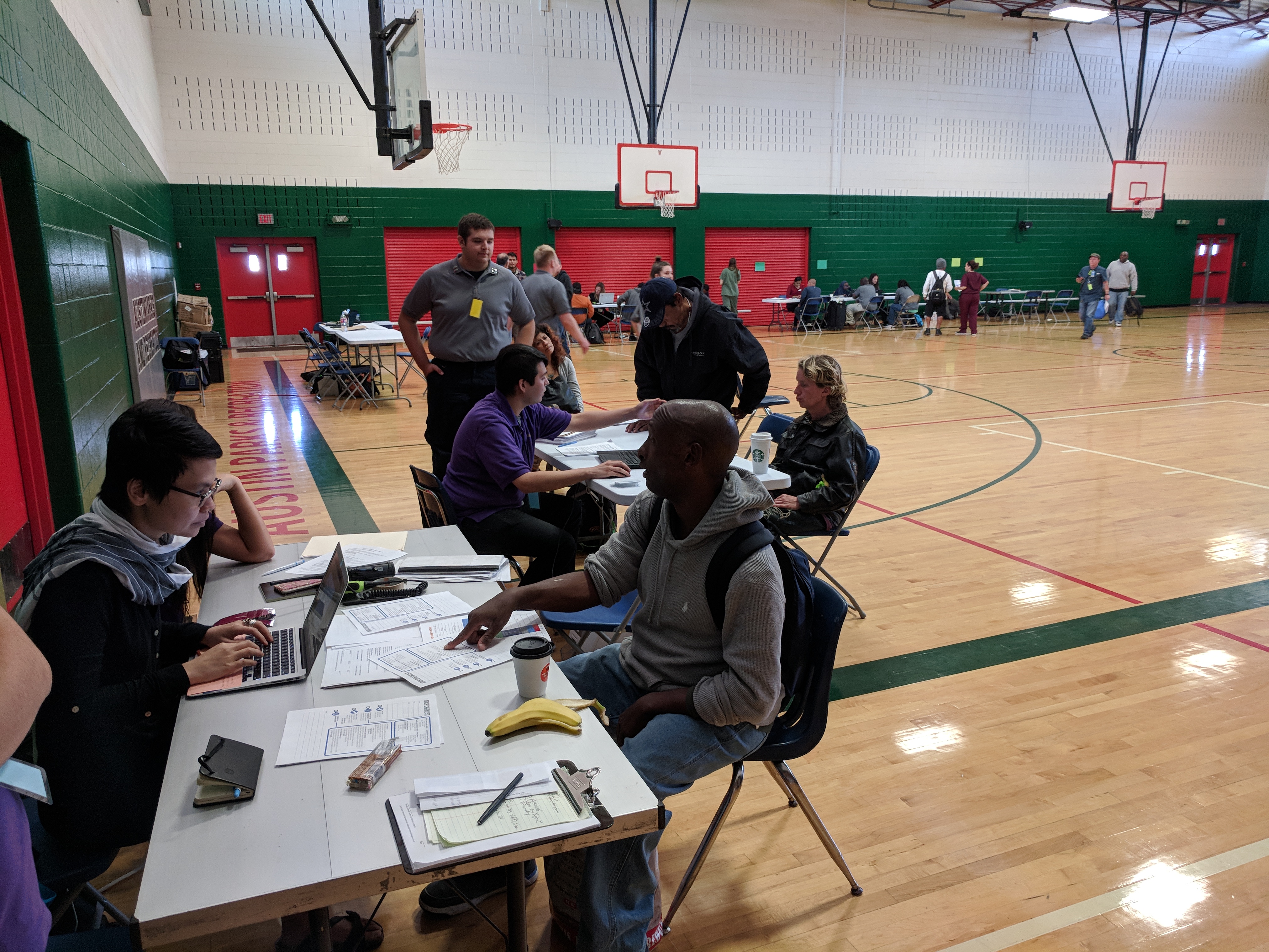 Tables are set up in the outer edges of a school gym. There are people sitting at each of these tables, using laptops and referencing papers.